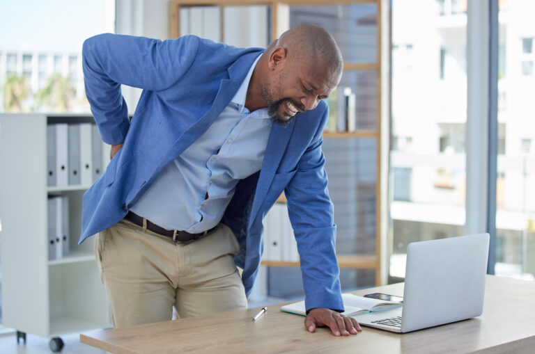 A man in a blue suit shows pain while resting one hand on a table and using the other to hold his lower back.